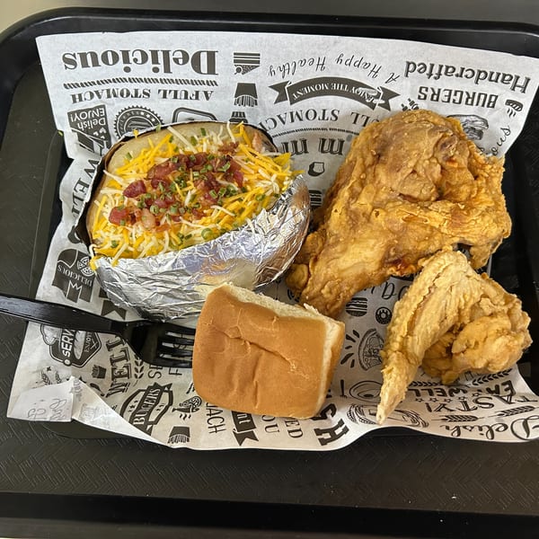 Fried chicken, a loaded baked potato in foil, and a dinner roll on a tray with patterned paper.