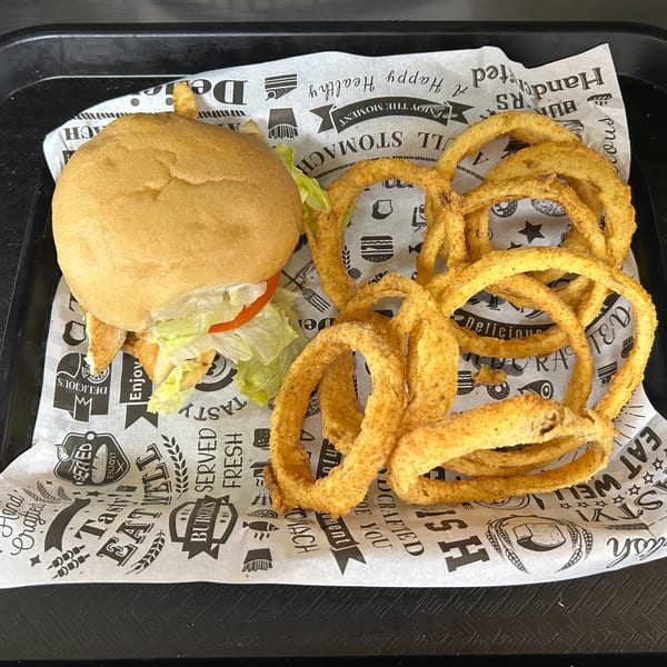 A chicken sandwich with lettuce and tomato, alongside a pile of golden onion rings on branded paper.